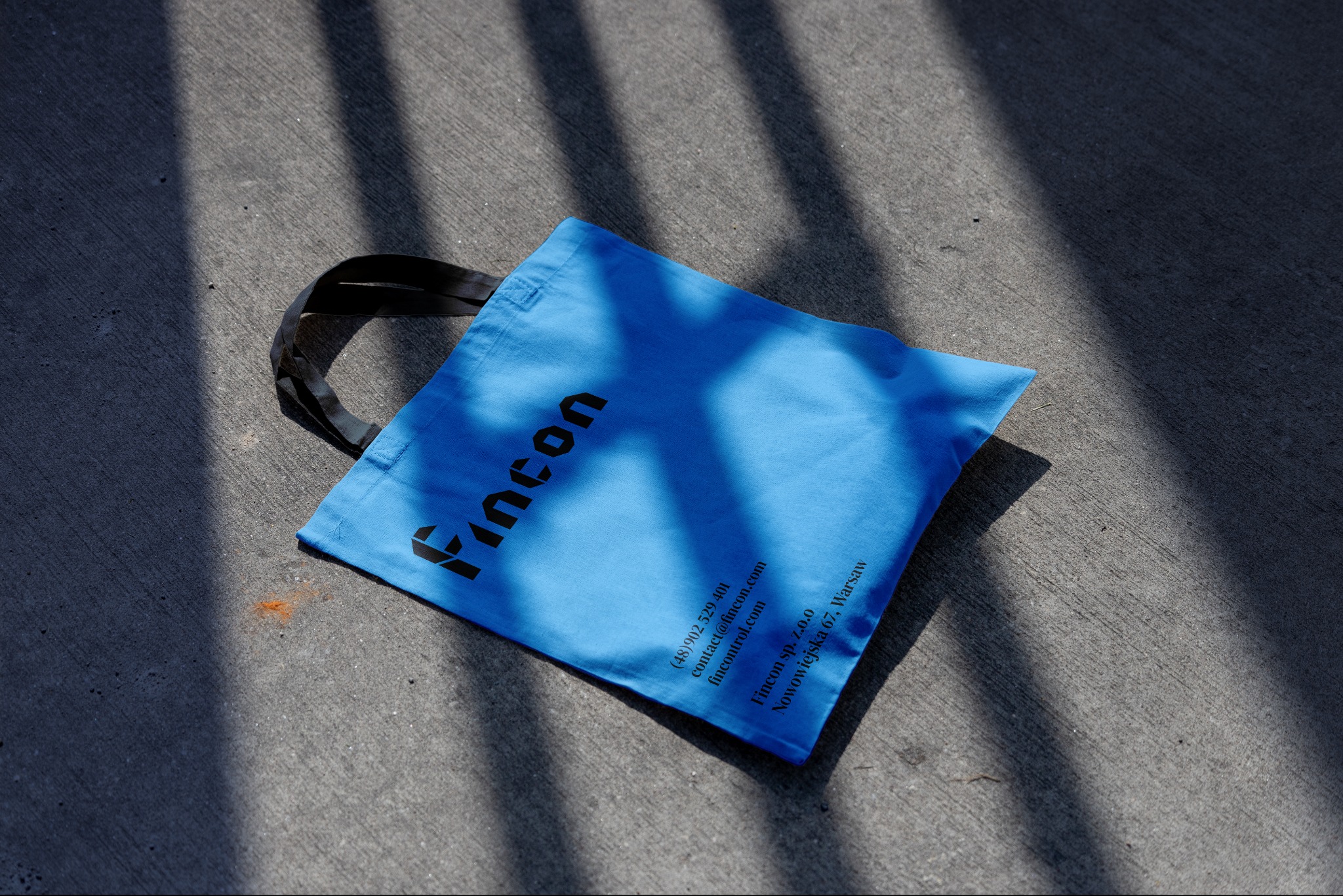Jasnoniebieska torba materiałowa z czarnym nadrukiem logo Fincon i danymi kontaktowymi leży na szarym betonie w cieniu drzewa.