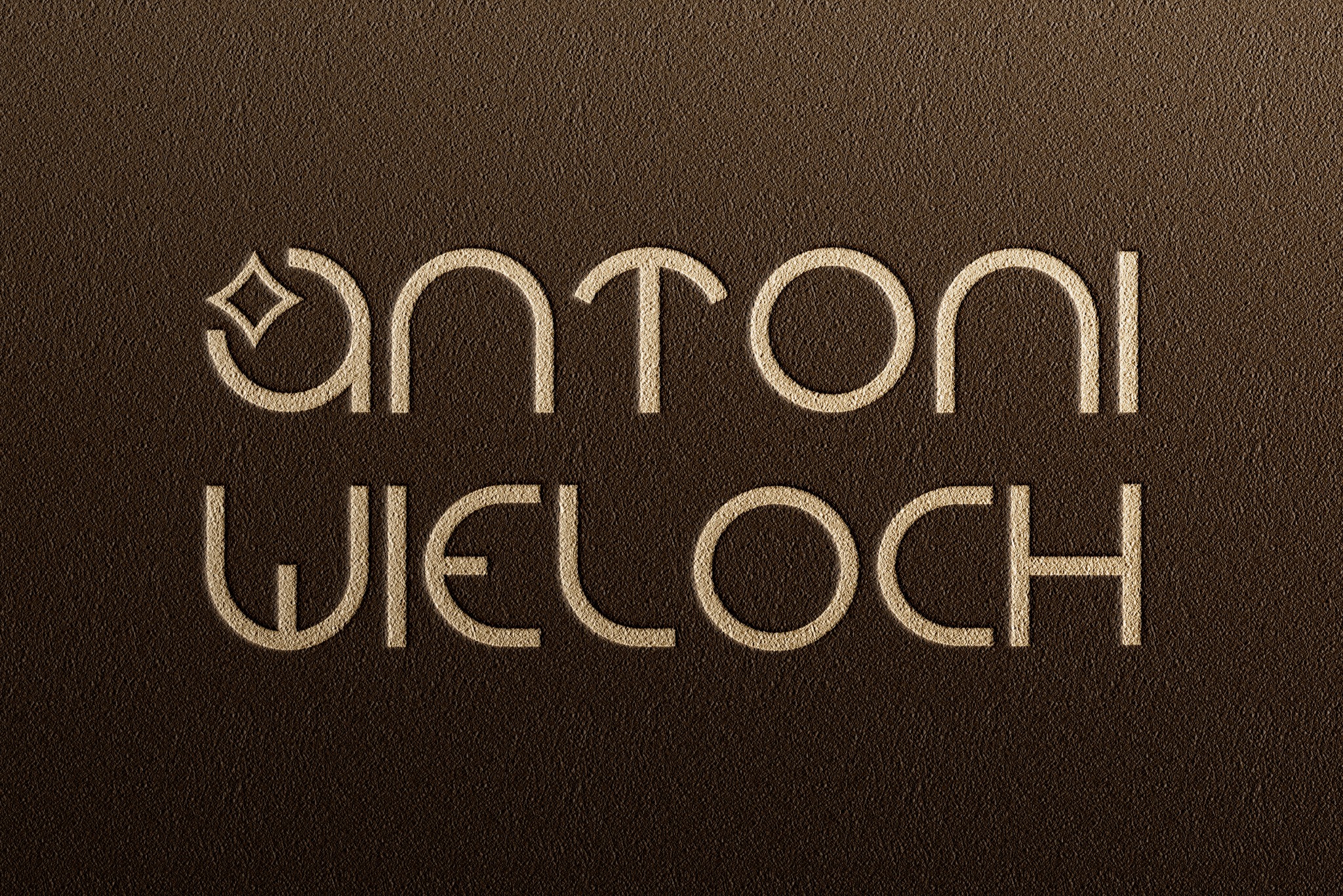 Złoty napis 'Antoni Wieloch' z elementem graficznym w miejscu litery 'a' na brązowym, teksturowanym tle.