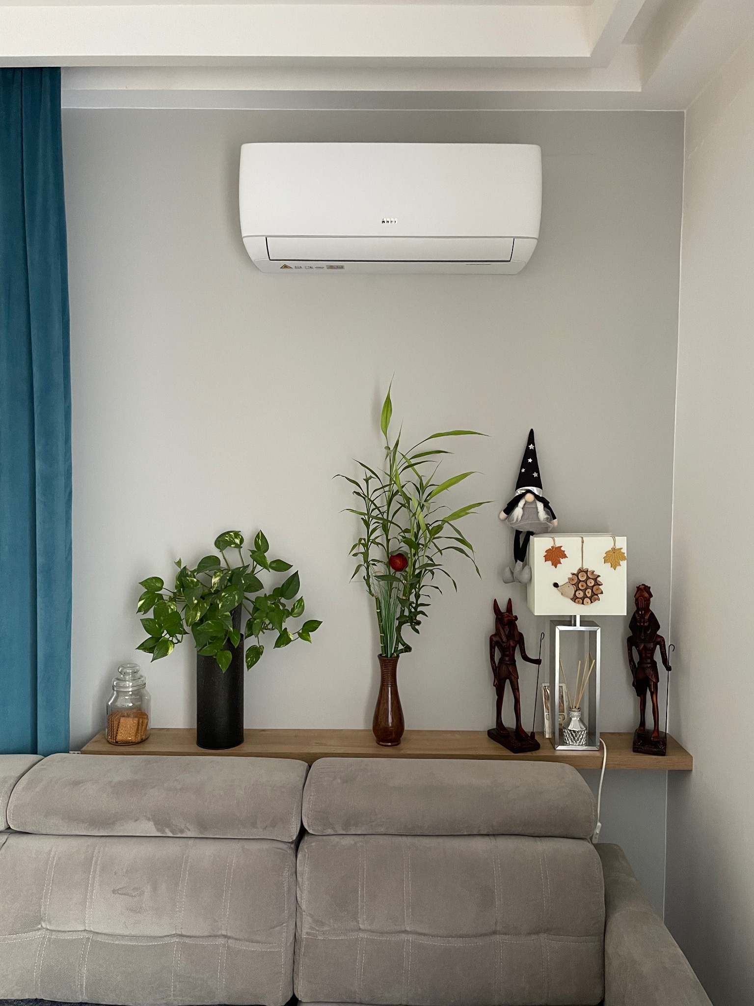 Biały klimatyzator ścienny zamontowany w salonie nad szarą sofą, z dekoracjami na półce, w tym rośliny, lampa z motywem jeża i figurki.