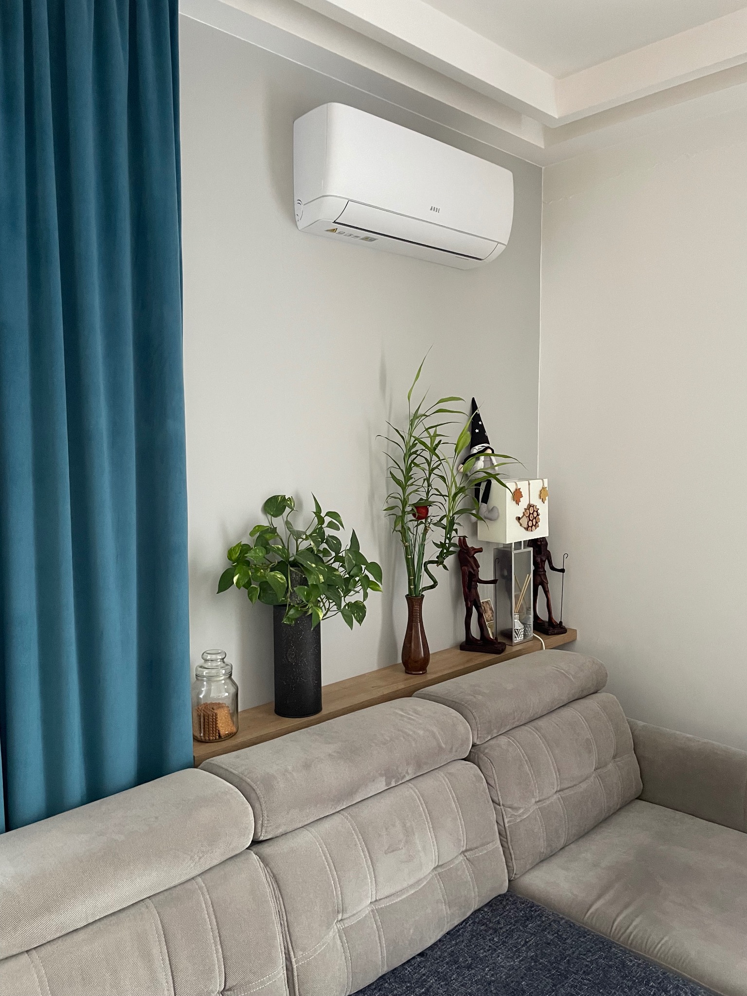 Biała jednostka klimatyzacyjna zamontowana nad szarą sofą w salonie, obok turkusowych zasłon i półki z dekoracjami.
