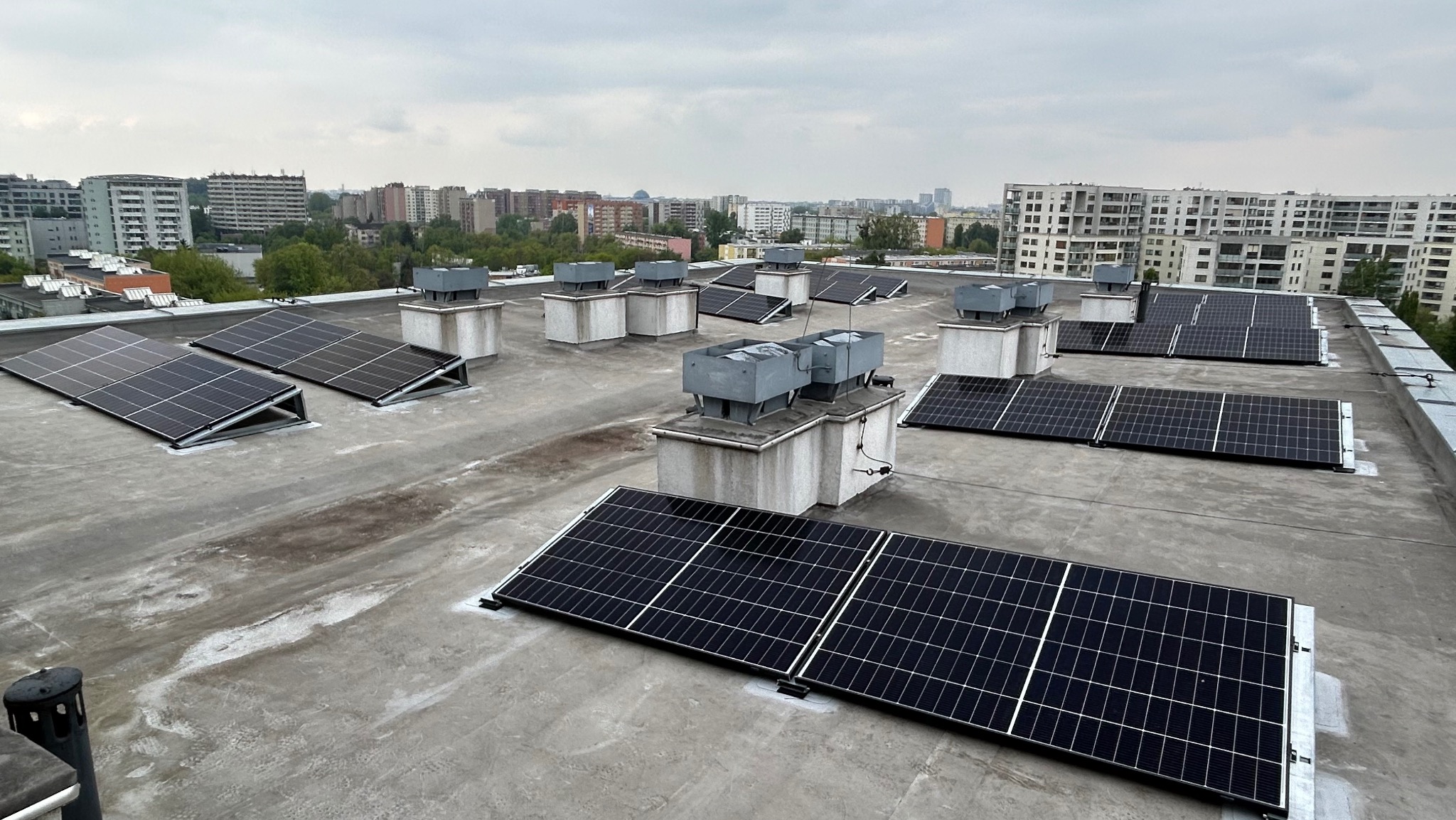Panele słoneczne zamontowane na płaskim dachu bloku mieszkalnego, konstrukcja zgrzewana do membrany dachowej, widok na panoramę miasta w tle.