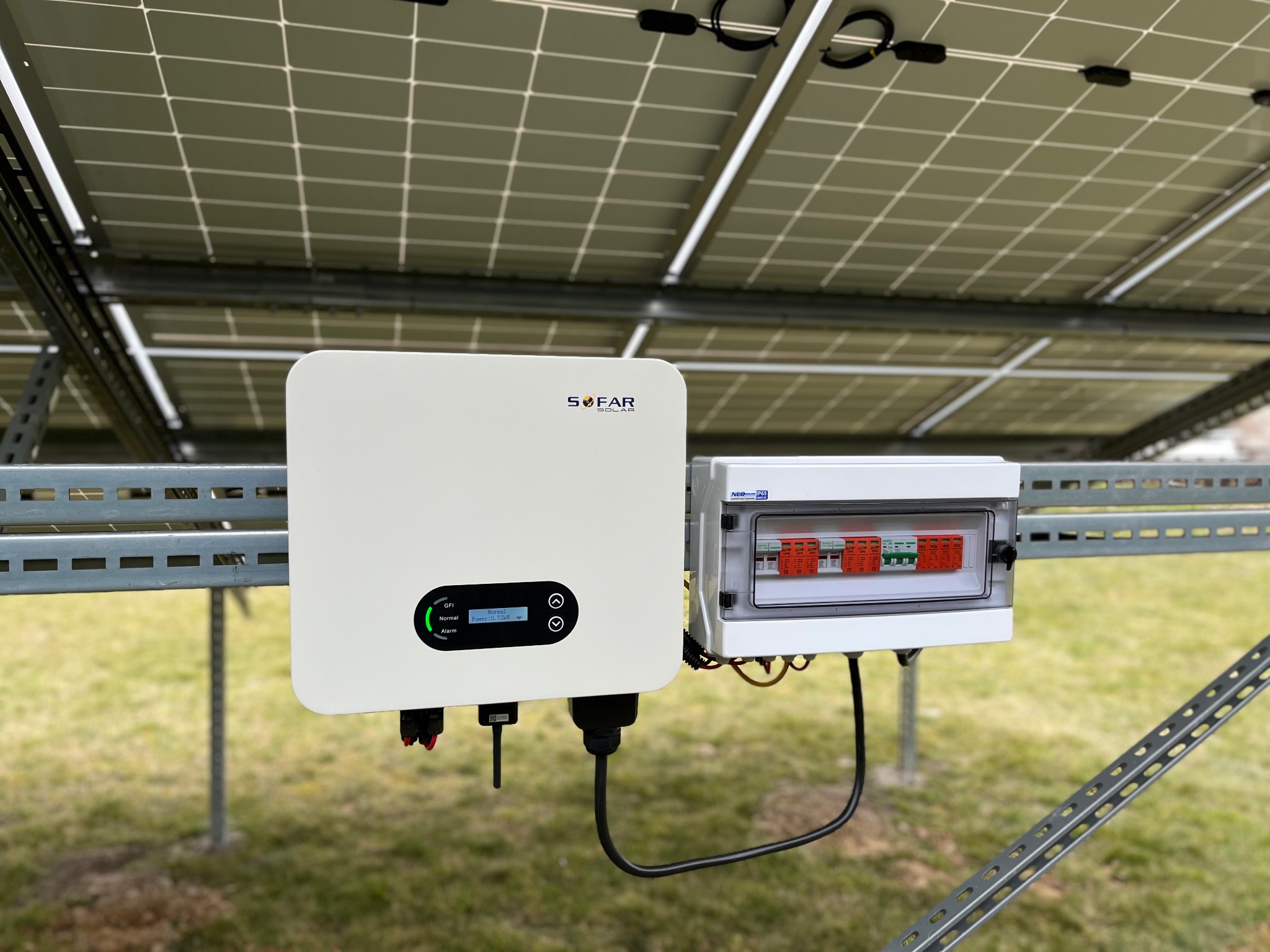 Biały inwerter Sofar Solar zamontowany na metalowej konstrukcji z panelami fotowoltaicznymi, obok skrzynka z bezpiecznikami, widoczne okablowanie i trawnik w tle.