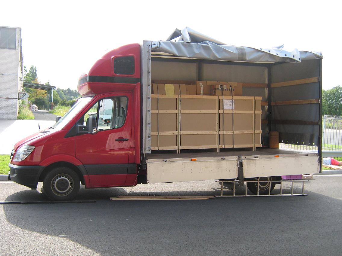 Czerwony samochód dostawczy z otwartą przestrzenią ładunkową, w której znajdują się duże, drewniane skrzynie zabezpieczone pasami. Z tyłu pojazdu widoczna drabina. Tło stanowią budynki i zieleń.