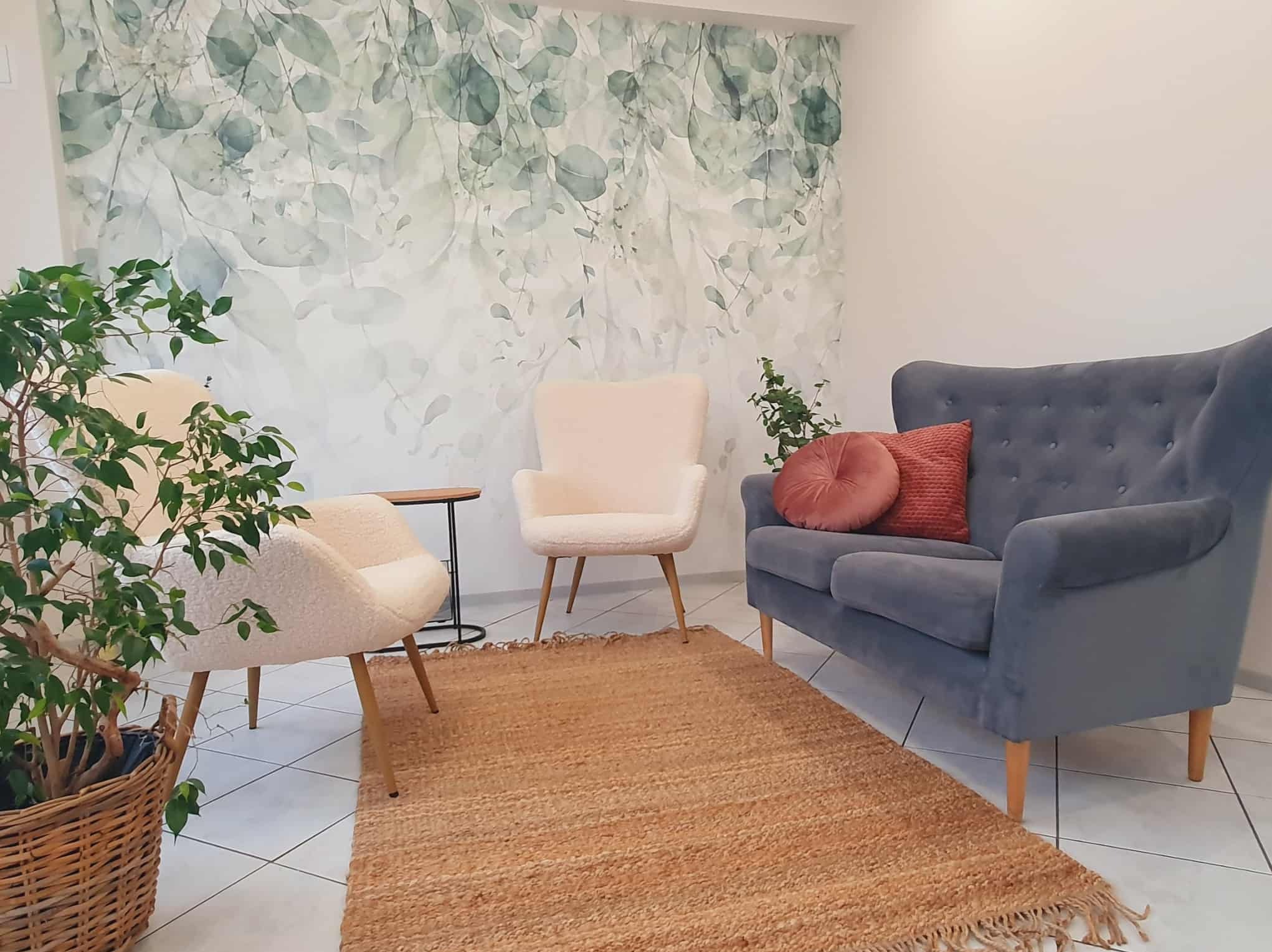 Jasne, przytulne wnętrze gabinetu z sofą, fotelami, dywanem i roślinami. Na ścianie tapeta w liście. Stonowana kolorystyka, relaksująca atmosfera.