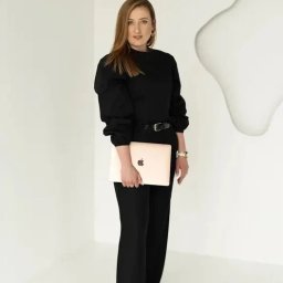 Zyskuj z Martą - Doradca Kredytowy Kraków - Kobieta w czarnym stroju trzyma laptopa na tle białego, minimalistycznego wnętrza. Profesjonalny wizerunek doradcy finansowego.