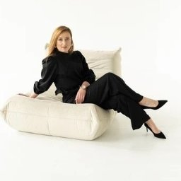 Zyskuj z Martą - Doradca Kredytowy Kraków - Kobieta w czarnym kombinezonie siedzi na beżowej pufie na białym tle. Widoczny zegarek na ręce i czarne szpilki. Minimalistyczna sesja w studio.