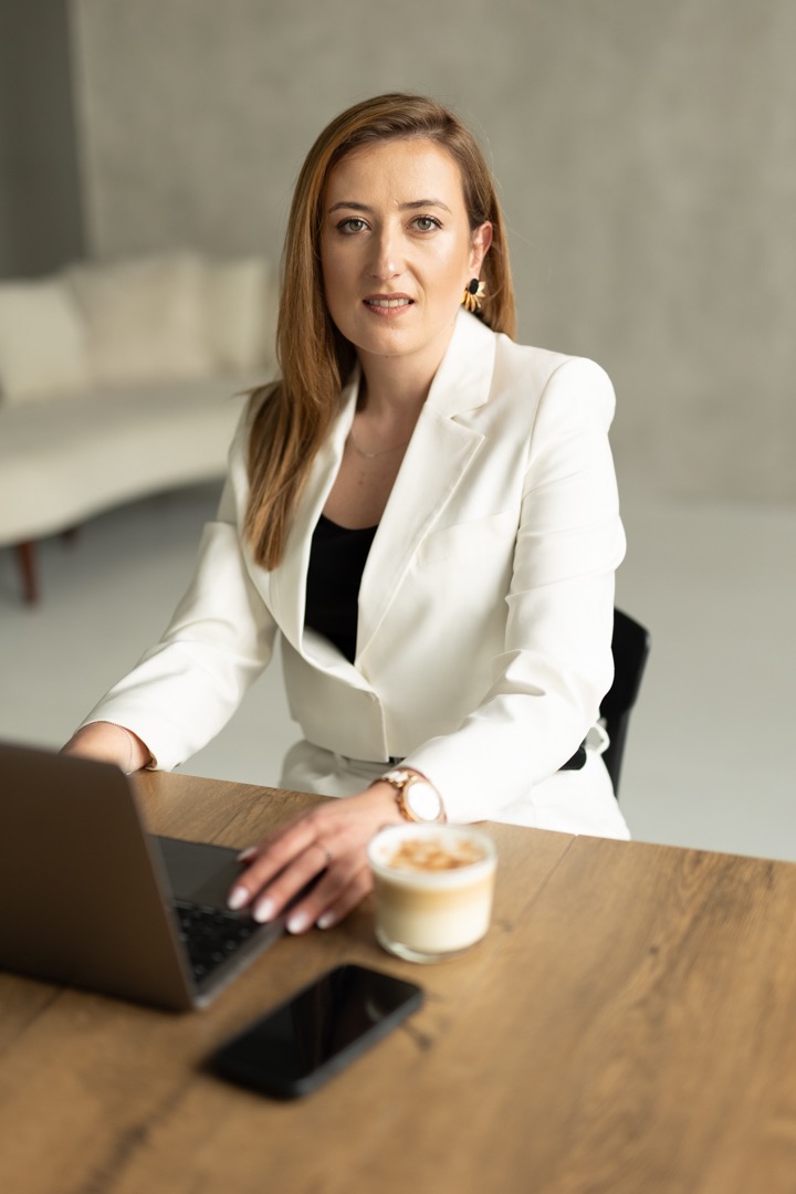 Kobieta w białym żakiecie pracuje na laptopie przy drewnianym biurku, obok stoi kawa latte i telefon.