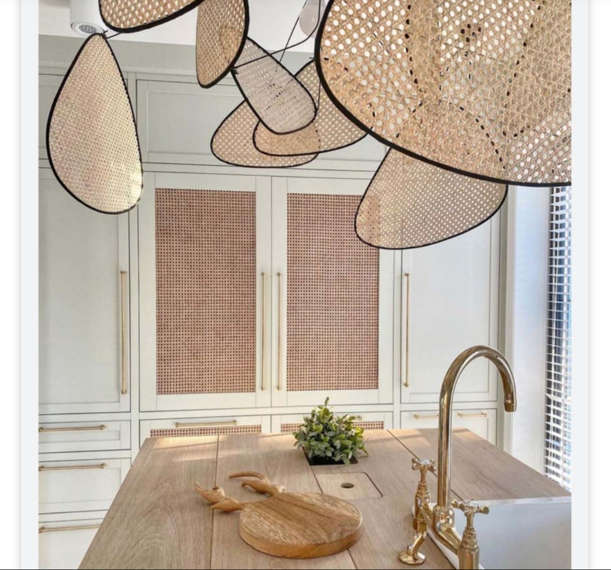 Jasna kuchnia z drewnianym blatem wyspowym, złotą armaturą i rattanowymi frontami szafek oraz dekoracyjną lampą sufitową z elementami rattanowymi.