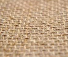 Zbliżenie na splot tkaniny jutowej o nieregularnej fakturze, widoczne włókna i prześwity, w ciepłej tonacji beżu i brązu, z rozmytym tłem.