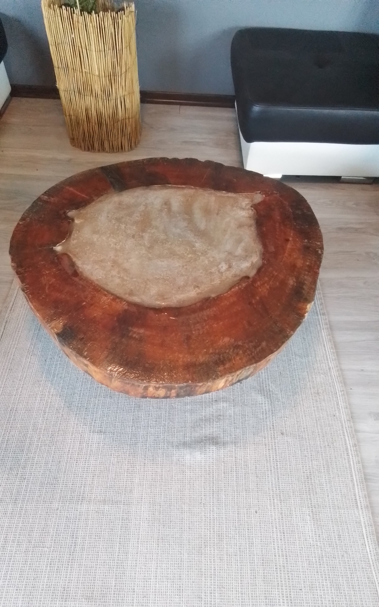 Niski stolik kawowy z plastra drewna z żywicznym wypełnieniem, ustawiony na jasnoszarym dywaniku, obok plecionego kosza i czarno-białego pufa.