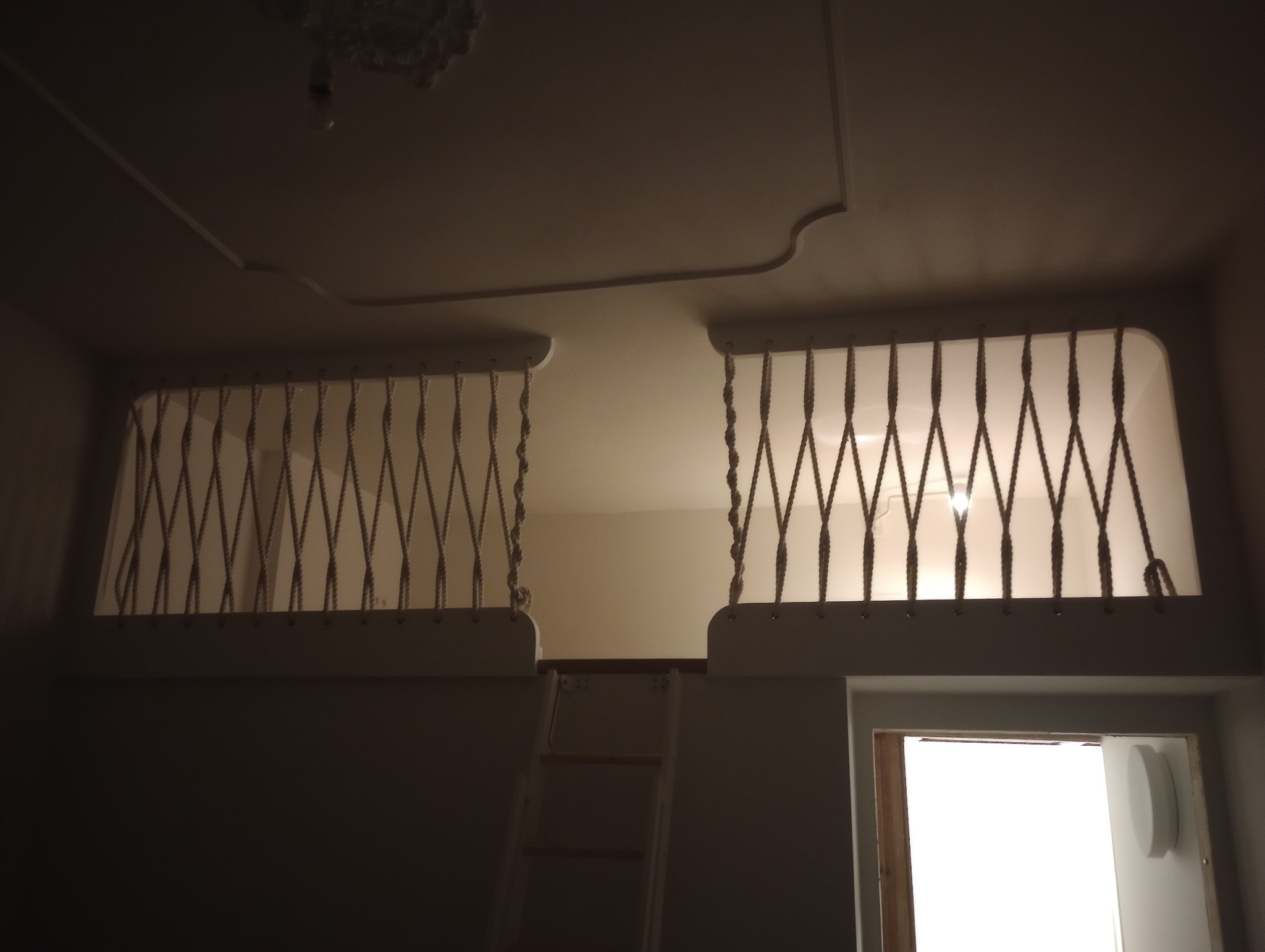 Wnętrze pokoju z antresolą zabezpieczoną białymi barierkami z grubych lin, widoczna drabina i fragment okna.