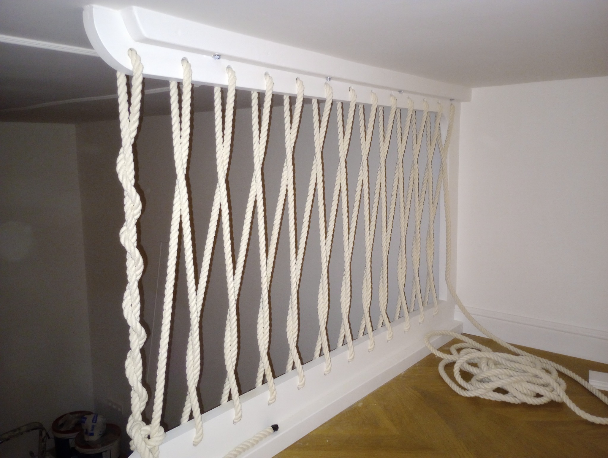 Biała balustrada schodowa wykonana z grubych, plecionych lin przymocowanych do białej ramy, widok z bliska, w tle jasne ściany i podłoga.