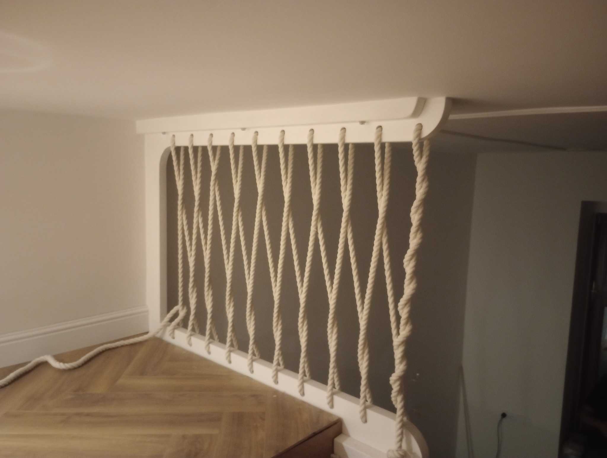 Barierka schodowa z białych drewnianych elementów i grubych, beżowych lin tworzących oryginalne wypełnienie, widok z dołu na drewnianą podłogę.