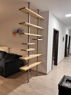 Regał z jasnego drewna i metalowych rur, pełniący funkcję ścianki działowej w nowoczesnym wnętrzu. Widoczne półki o nieregularnym rozmieszczeniu i czarna sofa w tle.