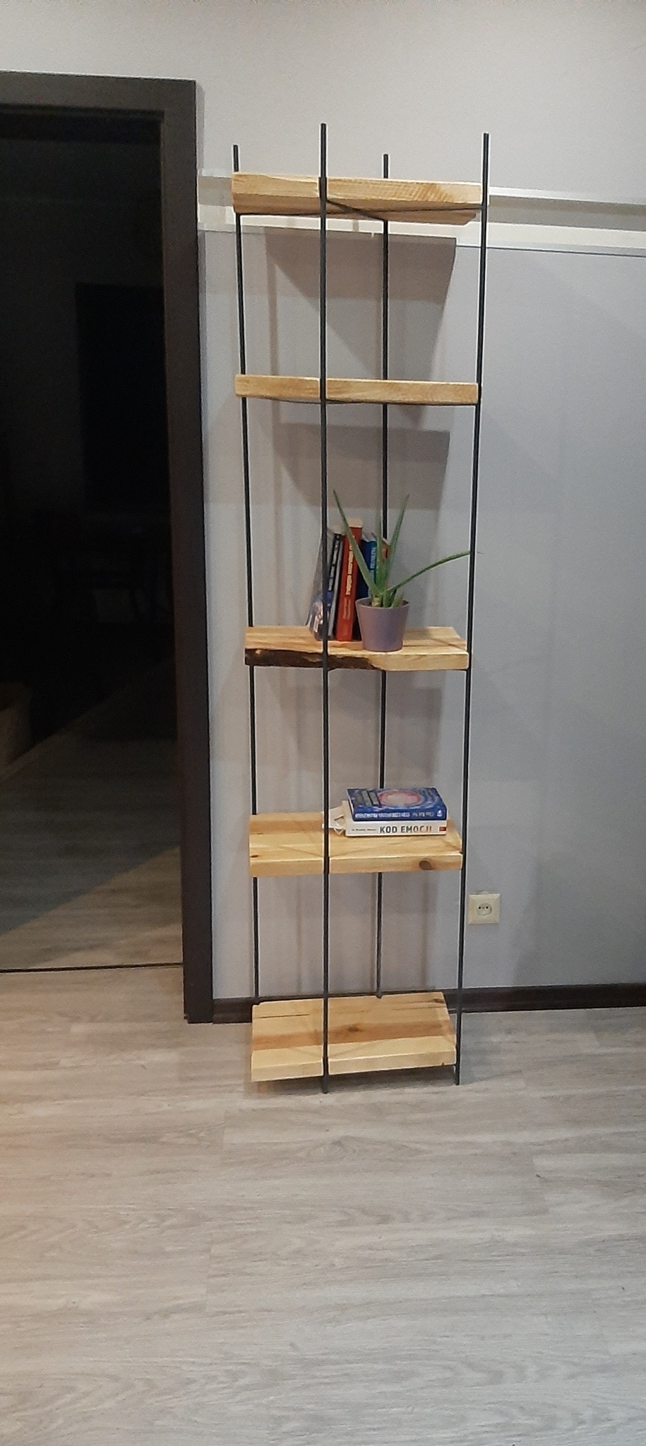Drewniana półka na książki z metalowymi prętami, na której stoją książki i mała roślina doniczkowa, ustawiona na tle szarej ściany i drewnianej podłogi.