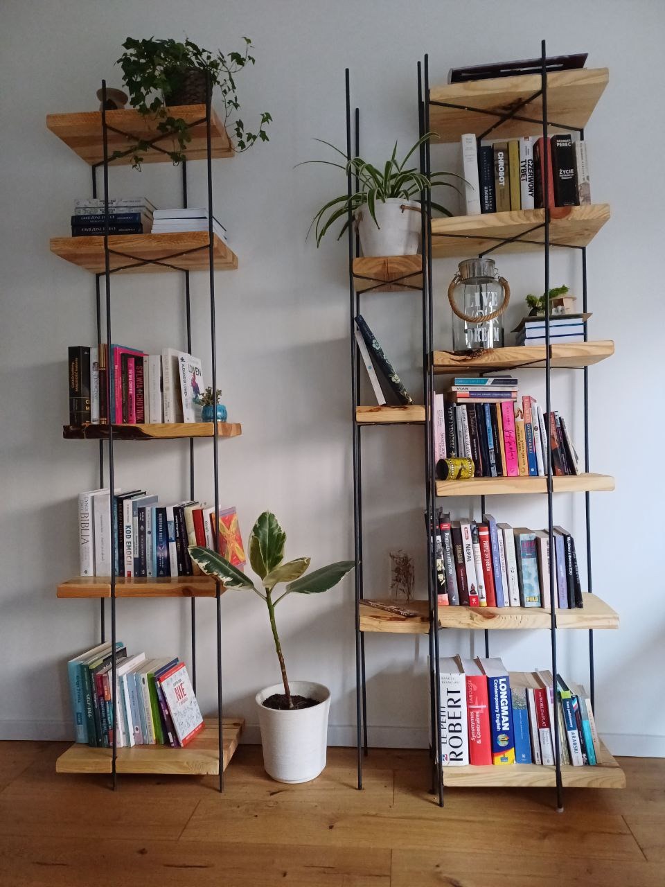 Dwie drewniane półki na książki z czarnymi metalowymi nogami, z książkami, roślinami doniczkowymi i dekoracjami, na drewnianej podłodze i białym tle ściany.
