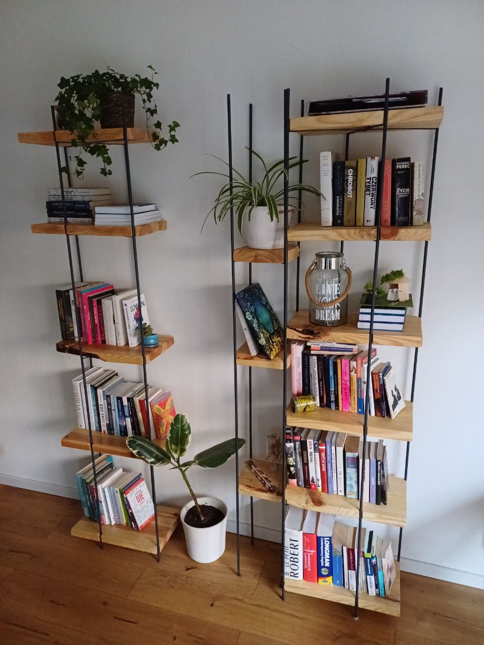Dwie otwarte półki na książki z drewna i metalu, wypełnione książkami, roślinami doniczkowymi i dekoracjami, stojące na drewnianej podłodze przy białej ścianie.