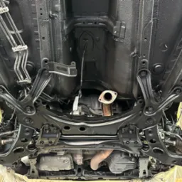 Podwozie samochodu po zabezpieczeniu antykorozyjnym, widoczne elementy zawieszenia i układu wydechowego pokryte czarną powłoką, z rdzawym fragmentem wydechu.