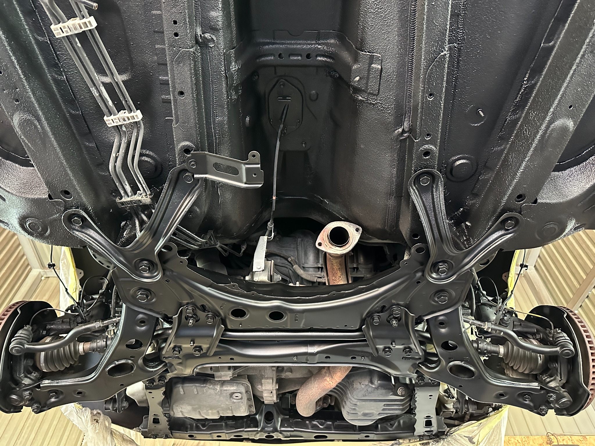 Podwozie samochodu po zabezpieczeniu antykorozyjnym, widoczne elementy zawieszenia i układu wydechowego pokryte czarną powłoką, z rdzawym fragmentem wydechu.