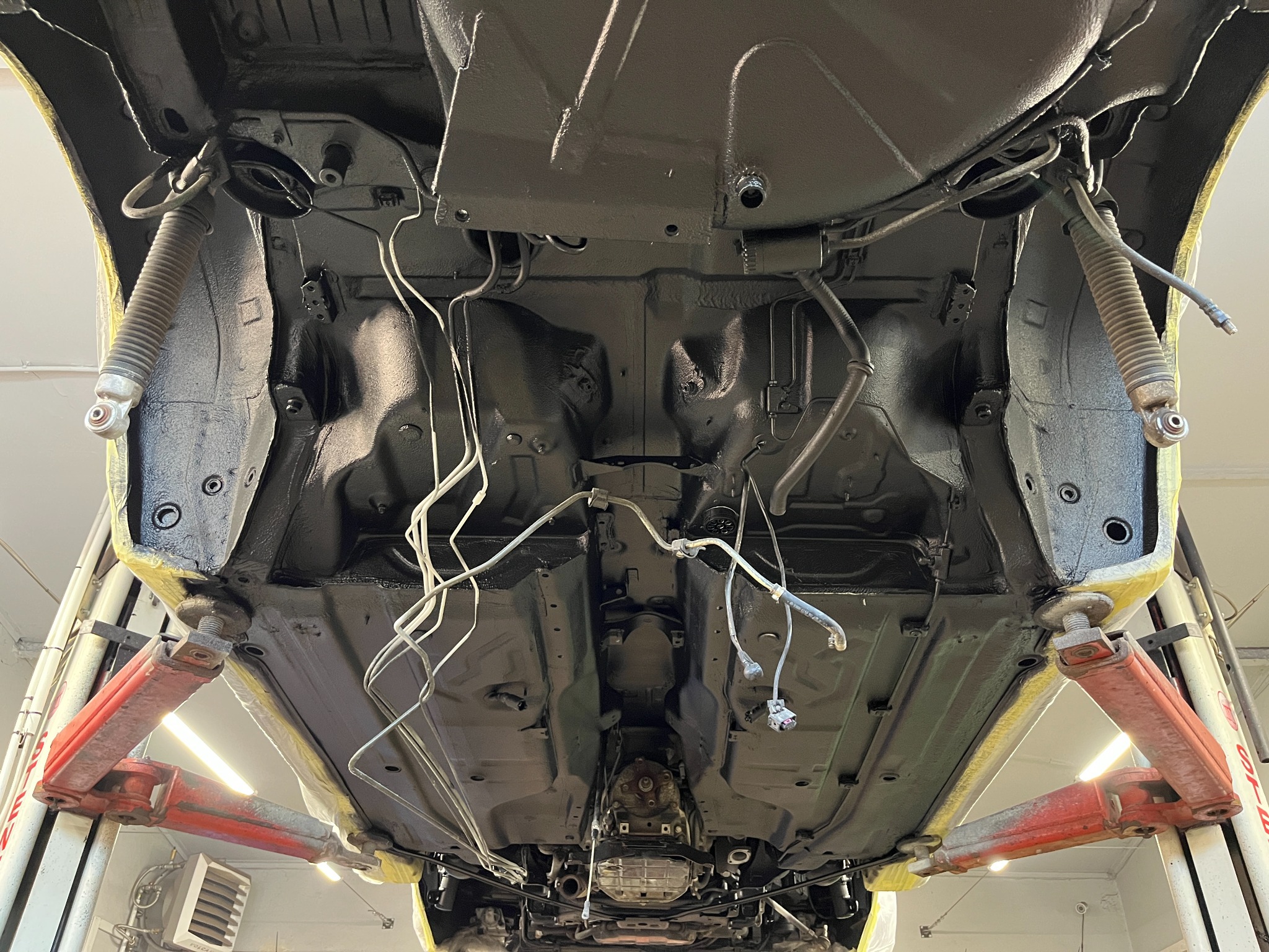 Podwozie samochodu na podnośniku, po zabezpieczeniu antykorozyjnym, widoczne elementy zawieszenia i przewody, żółta taśma maskująca na krawędziach.