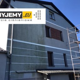 myjemy.eu - Dwupiętrowy dom z szarą elewacją, częściowo umytą ciśnieniowo, z widoczną różnicą między czystą a zabrudzoną powierzchnią. Obok ściany ustawione rusztowanie. W lewym górnym rogu widoczne logo firmy.
