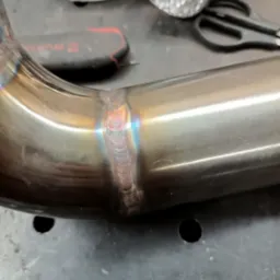 Metalowa rura z precyzyjnym spoinem, widoczne tęczowe przebarwienia termiczne, narzędzia w tle.