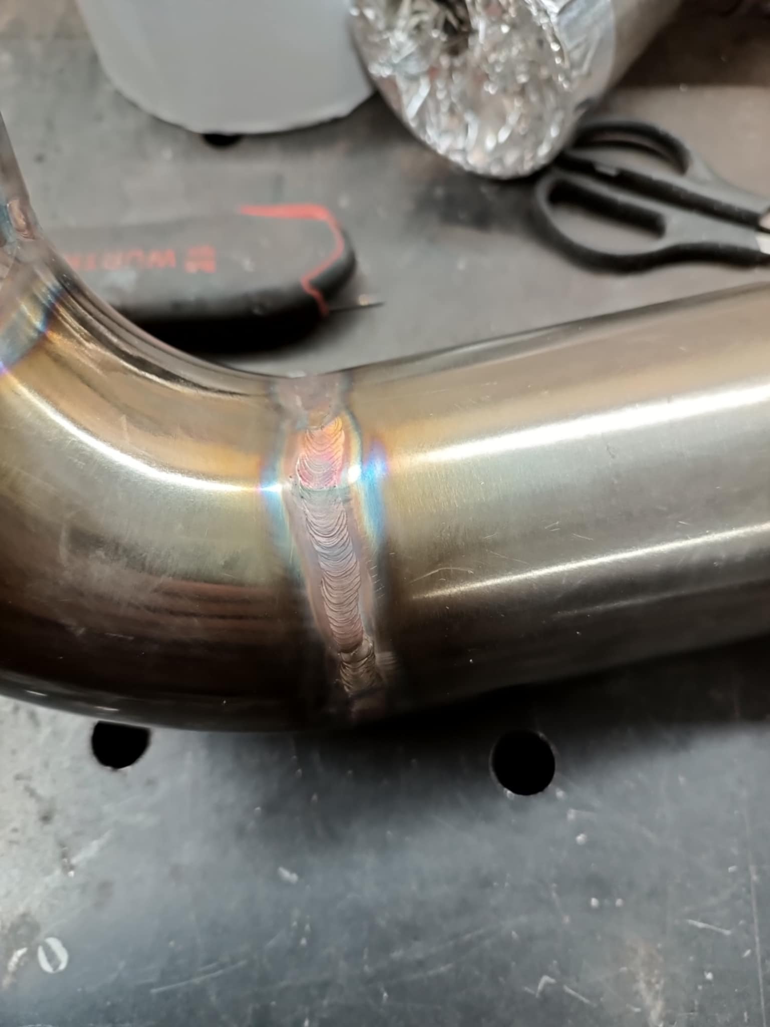 Metalowa rura z precyzyjnym spoinem, widoczne tęczowe przebarwienia termiczne, narzędzia w tle.