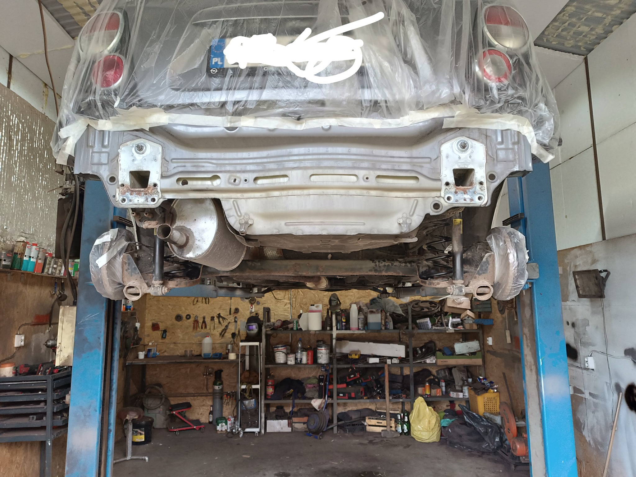 Samochód na podnośniku w warsztacie, zabezpieczony folią, widoczny tylny zderzak w trakcie naprawy lub przygotowania do lakierowania, na ścianach warsztatu narzędzia i akcesoria.