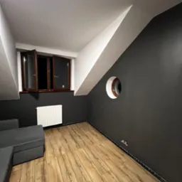Pokój na poddaszu z drewnianą podłogą i oknem. Ściany pomalowane na szaro, sufit biały. Nietypowe okrągłe okno w ścianie. Widoczny fragment sofy.