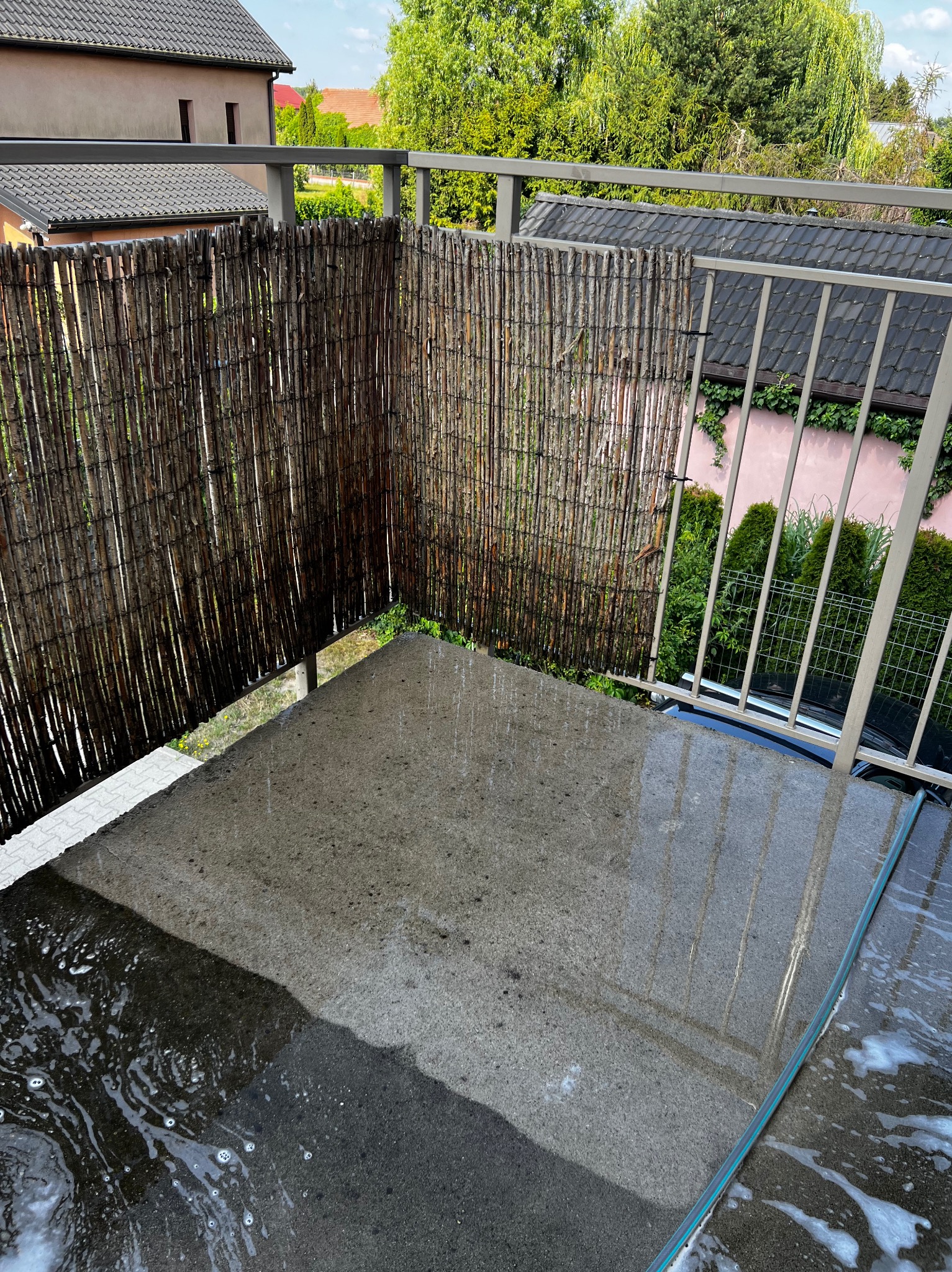Mokry, betonowy balkon po umyciu, woda z pianą spływa po powierzchni, widoczne bambusowe osłony i metalowe barierki, w tle zieleń i zabudowa.