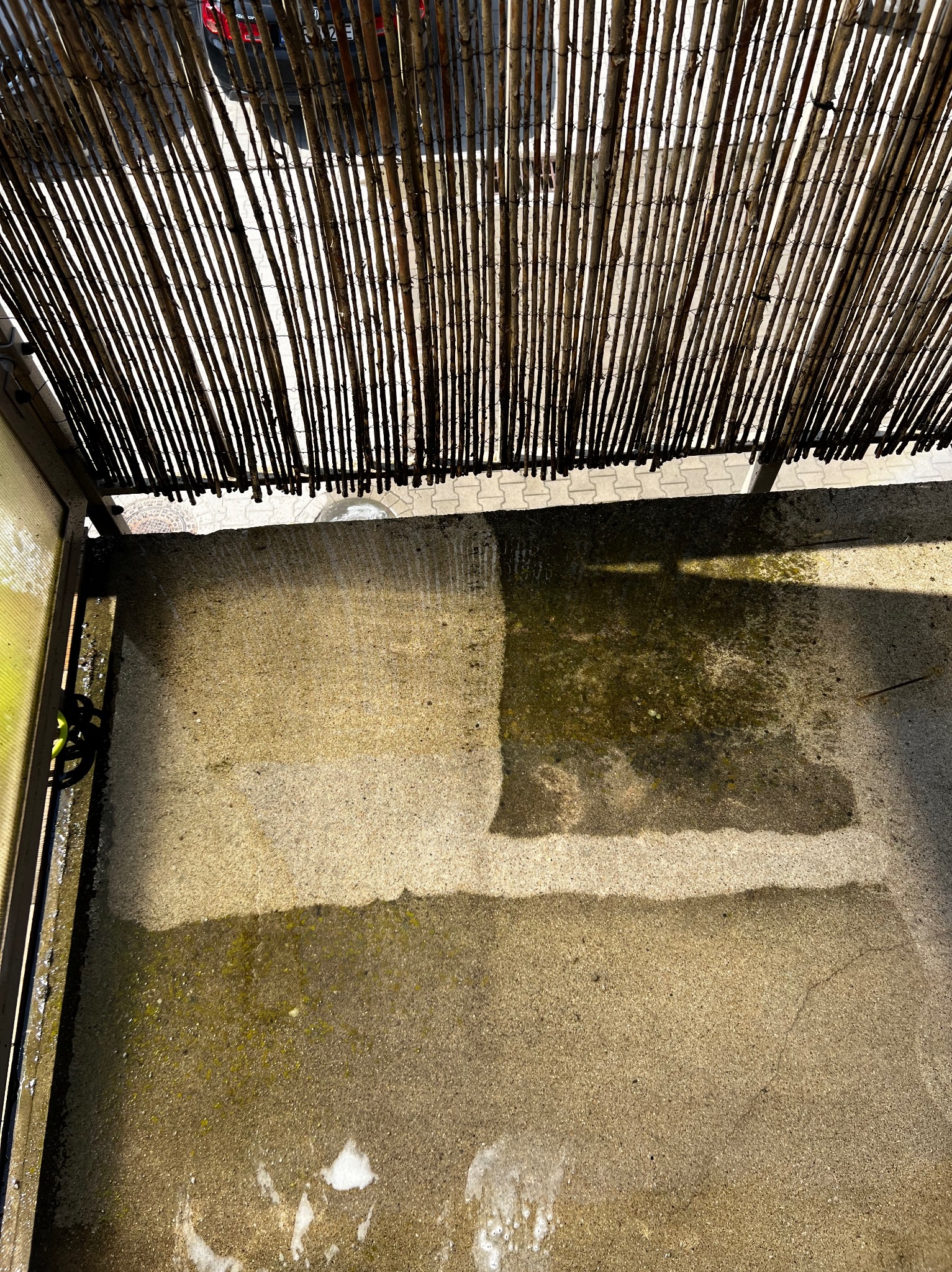 Fragment betonowego tarasu w Kiełczowie, częściowo umytego, z widoczną różnicą w kolorze i czystości, zadaszonego bambusową roletą.