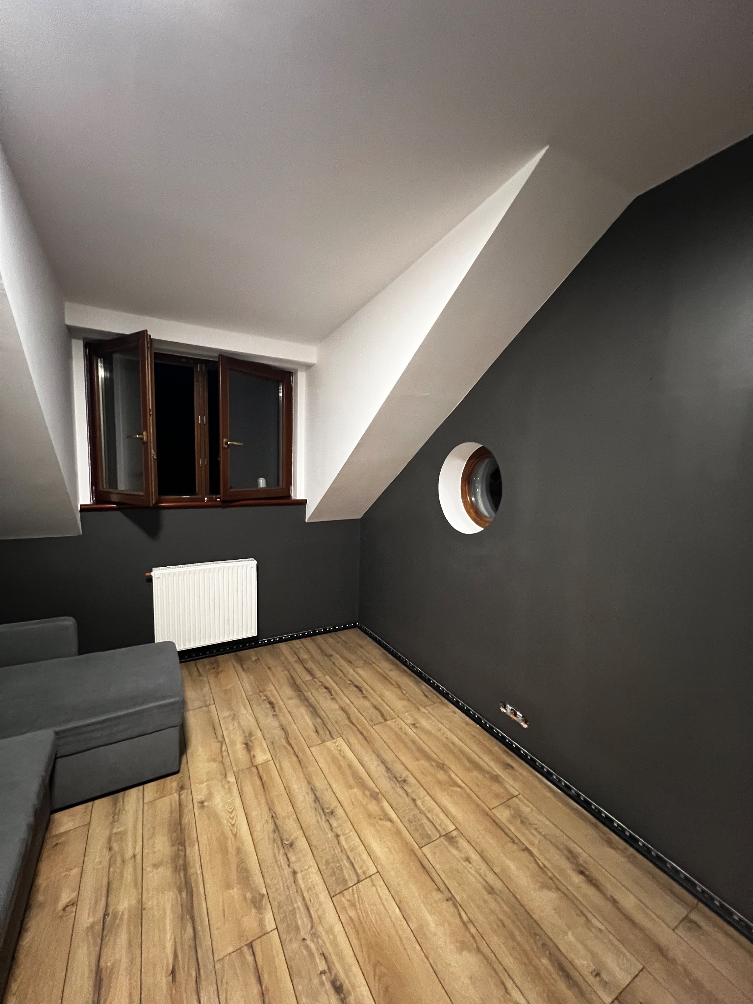 Pokój na poddaszu z drewnianą podłogą i oknem. Ściany pomalowane na szaro, sufit biały. Nietypowe okrągłe okno w ścianie. Widoczny fragment sofy.