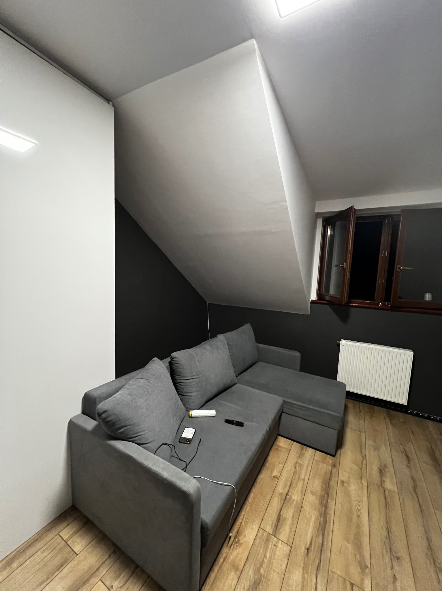 Szare panele podłogowe w pokoju na poddaszu z szarą kanapą i czarną ścianą. Widoczne otwarte okno i grzejnik. Oświetlenie sufitowe.