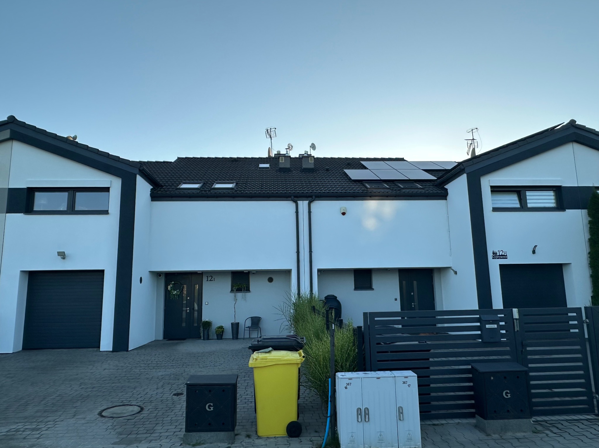 Bliźniak z białą elewacją, czarną stolarką i panelami słonecznymi na dachu. Widoczne kosze na śmieci i skrzynki elektryczne przed posesją. Płytki chodnikowe prowadzą do garażu.
