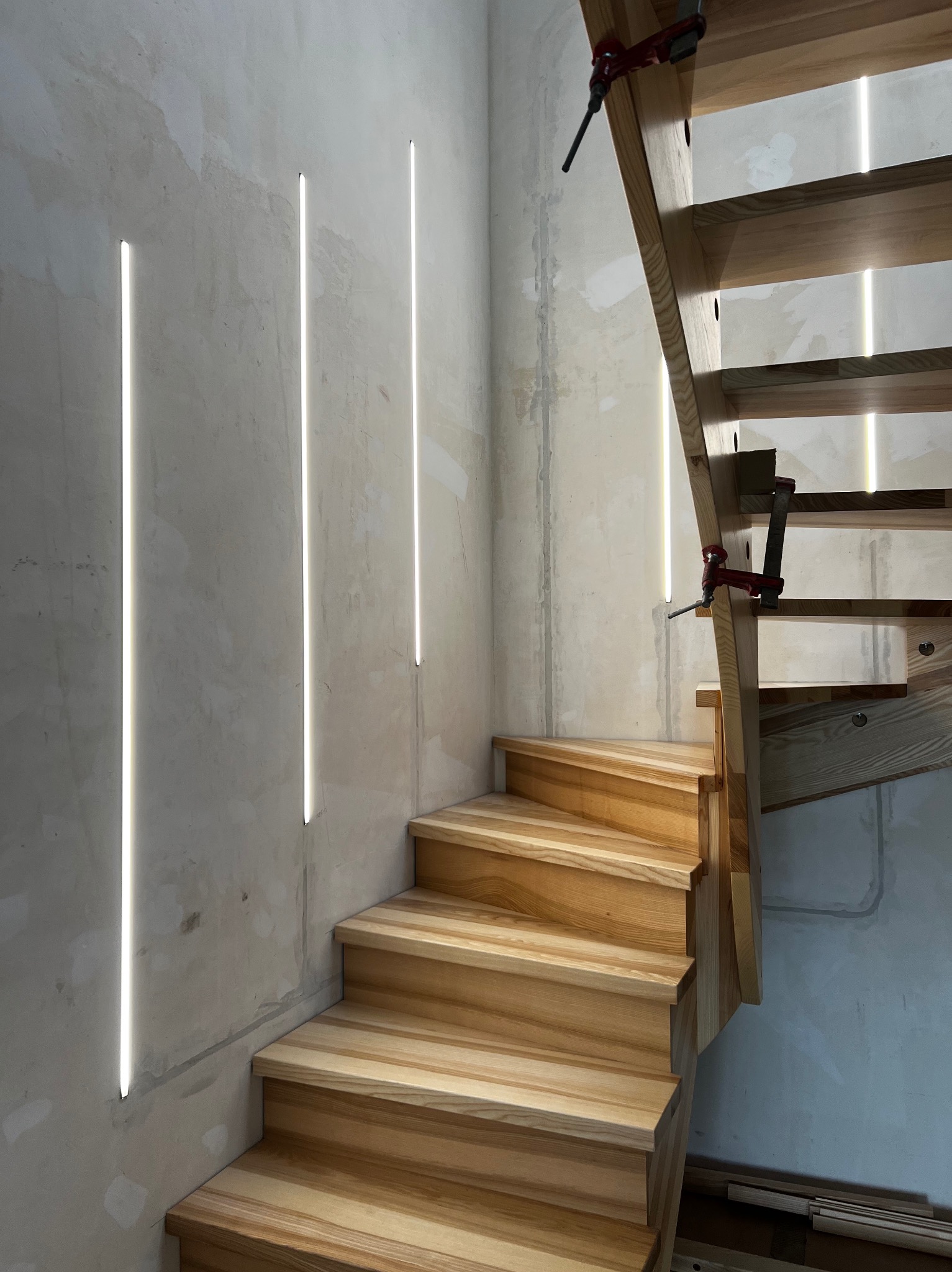 Drewniane schody w trakcie montażu, z widocznymi klamrami stolarskimi. Na ścianie pionowe, minimalistyczne oświetlenie LED. Surowy beton na ścianie.