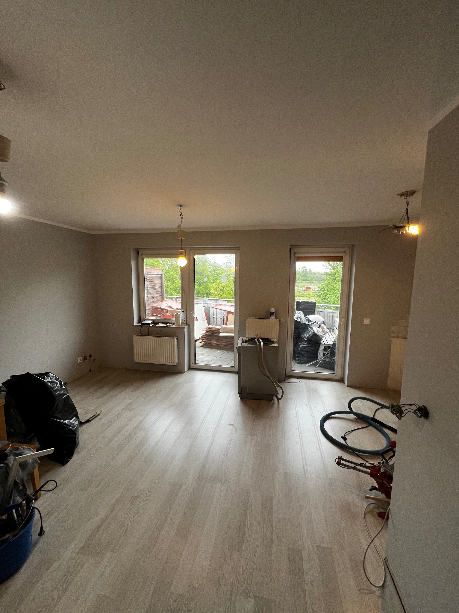 Jasne panele podłogowe w trakcie remontu mieszkania z widokiem na balkon, worki na śmieci, narzędzia i odkurzacz budowlany. Szare ściany i niedokończone oświetlenie.