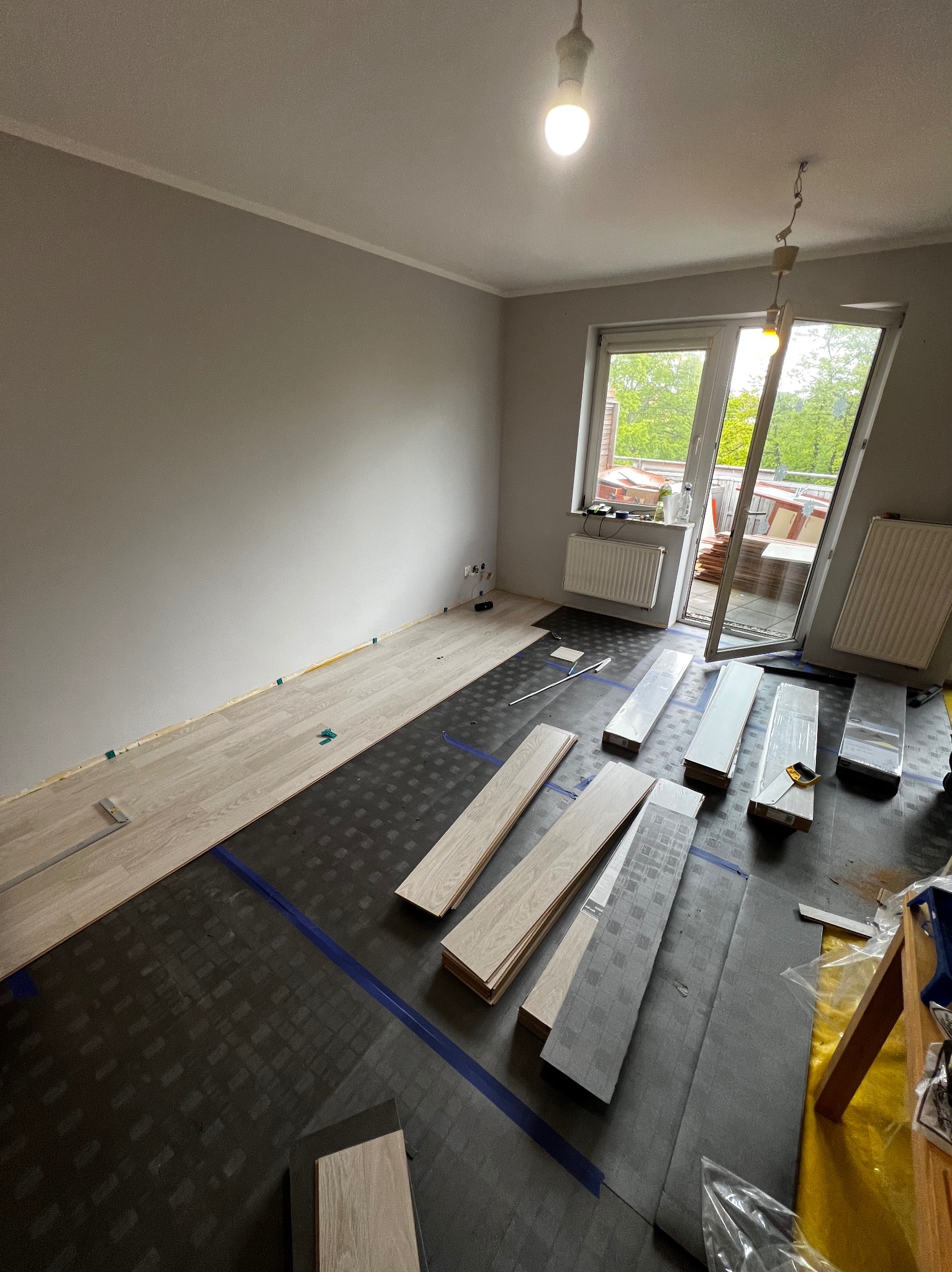 Układanie paneli podłogowych w pokoju z balkonem. Częściowo ułożona podłoga, widoczne panele, podkład i narzędzia. Ściany szare, pokój jasny.