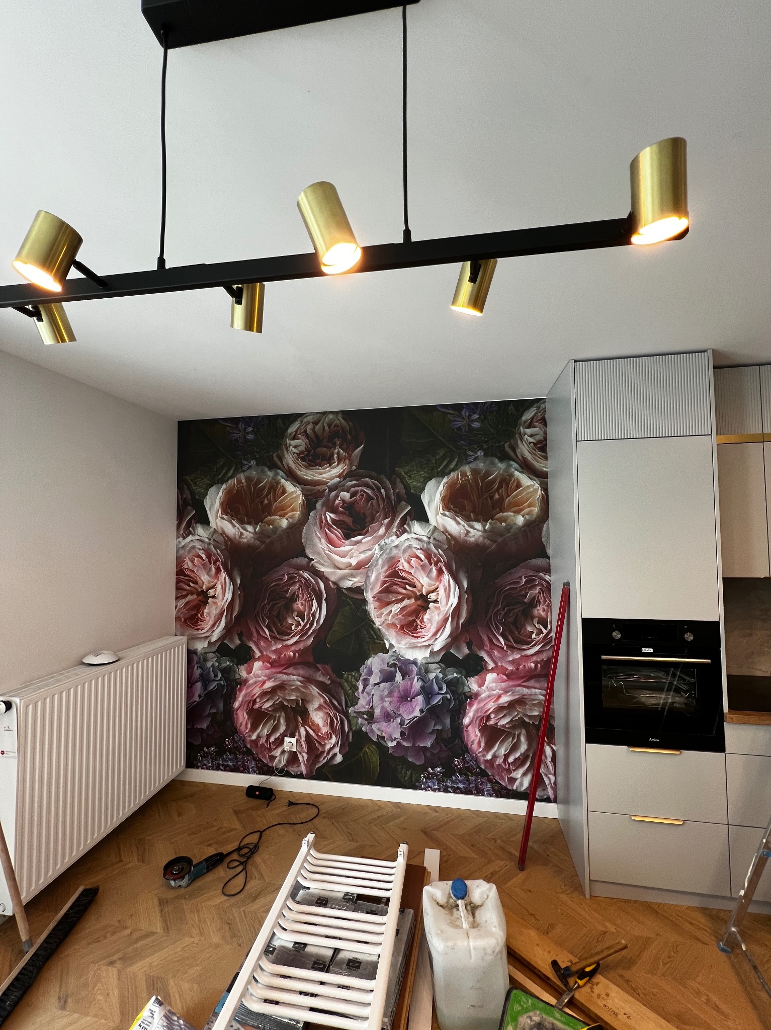 Kuchenne wnętrze w trakcie odświeżania: ściana z fototapetą w kwiaty, nowoczesne oświetlenie sufitowe i parkiet w jodełkę. Widoczne narzędzia i materiały budowlane.