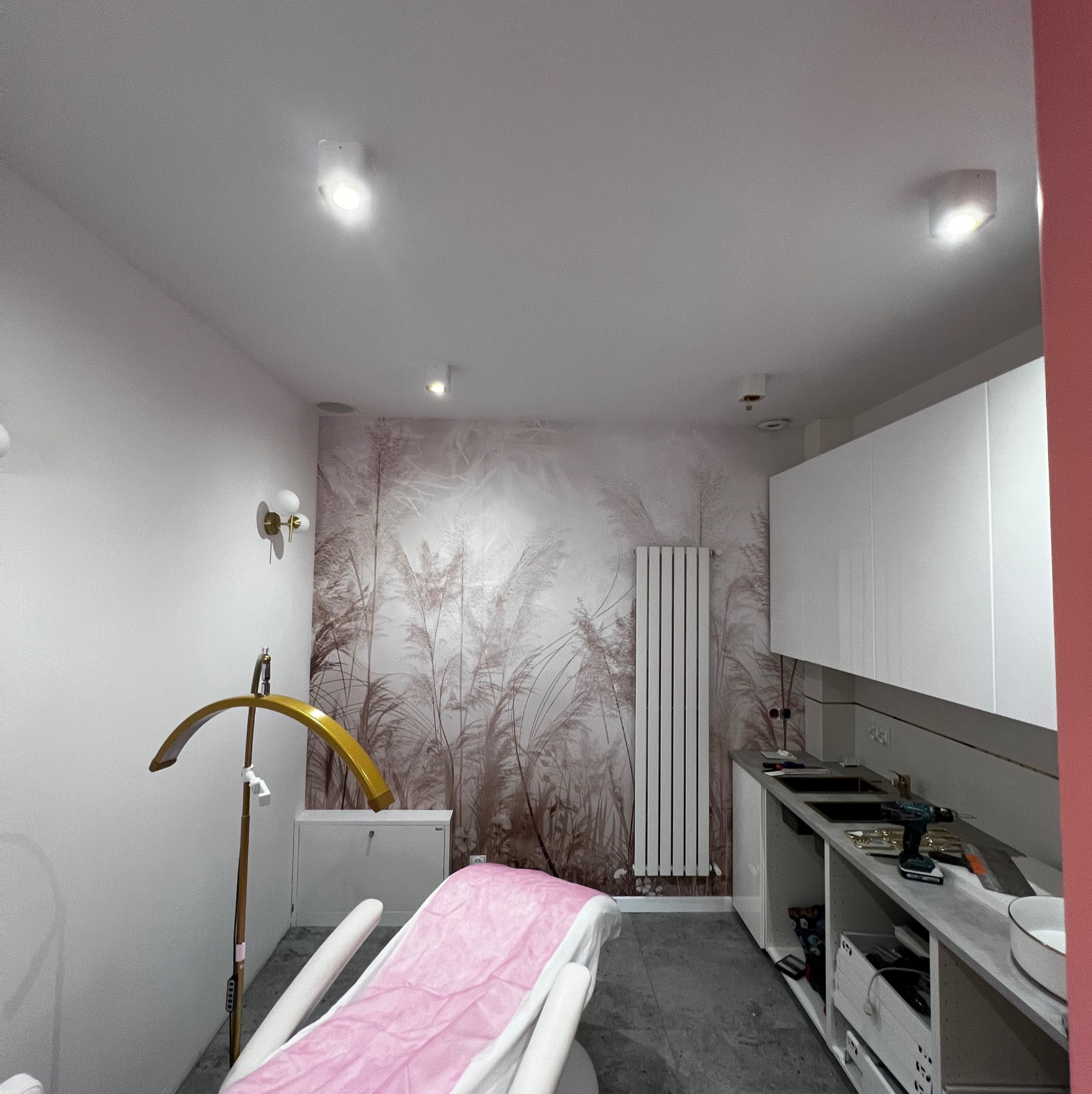 Nowoczesne wnętrze salonu kosmetycznego: fotel z różowym obiciem, lampa, szafki i ściana z motywem roślinnym. W tle widoczny grzejnik i narzędzia.