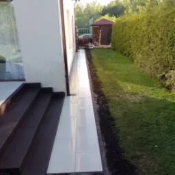 Ułożona, jasnoszara kostka brukowa tworząca ścieżkę wzdłuż ściany domu, z ciemnymi schodami wejściowymi i trawnikiem z żywopłotem w tle.