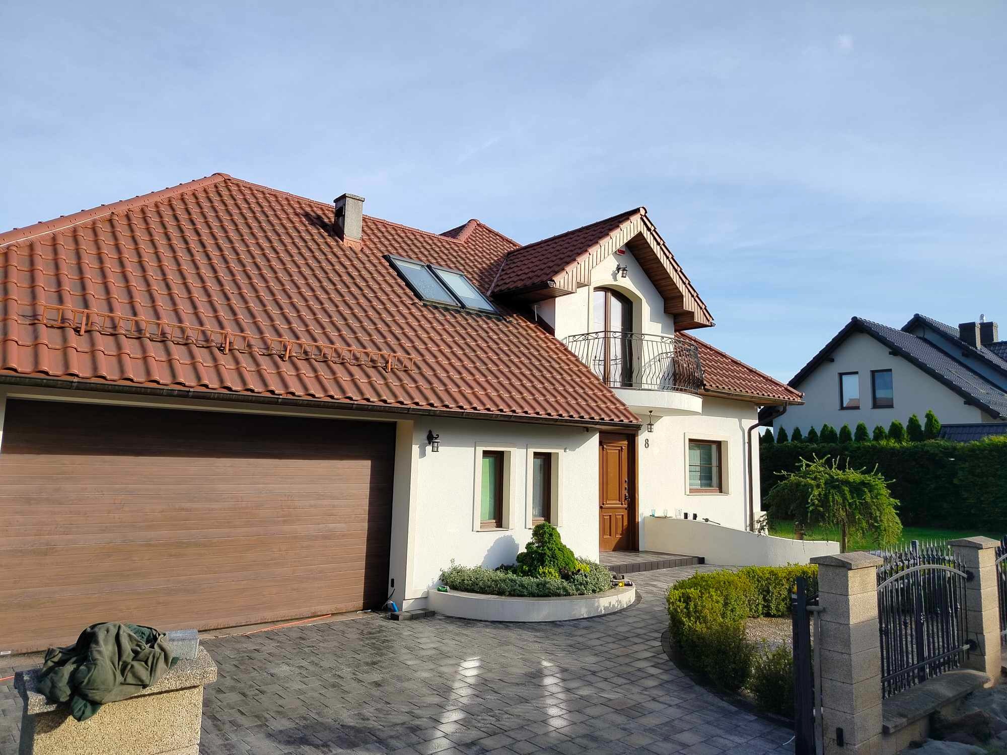 Nowoczesny dom jednorodzinny z brązową dachówką i garażem, otoczony kostką brukową ułożoną w jodełkę, widoczny fragment ogrodzenia z metalowymi przęsłami i kamiennymi słupkami.