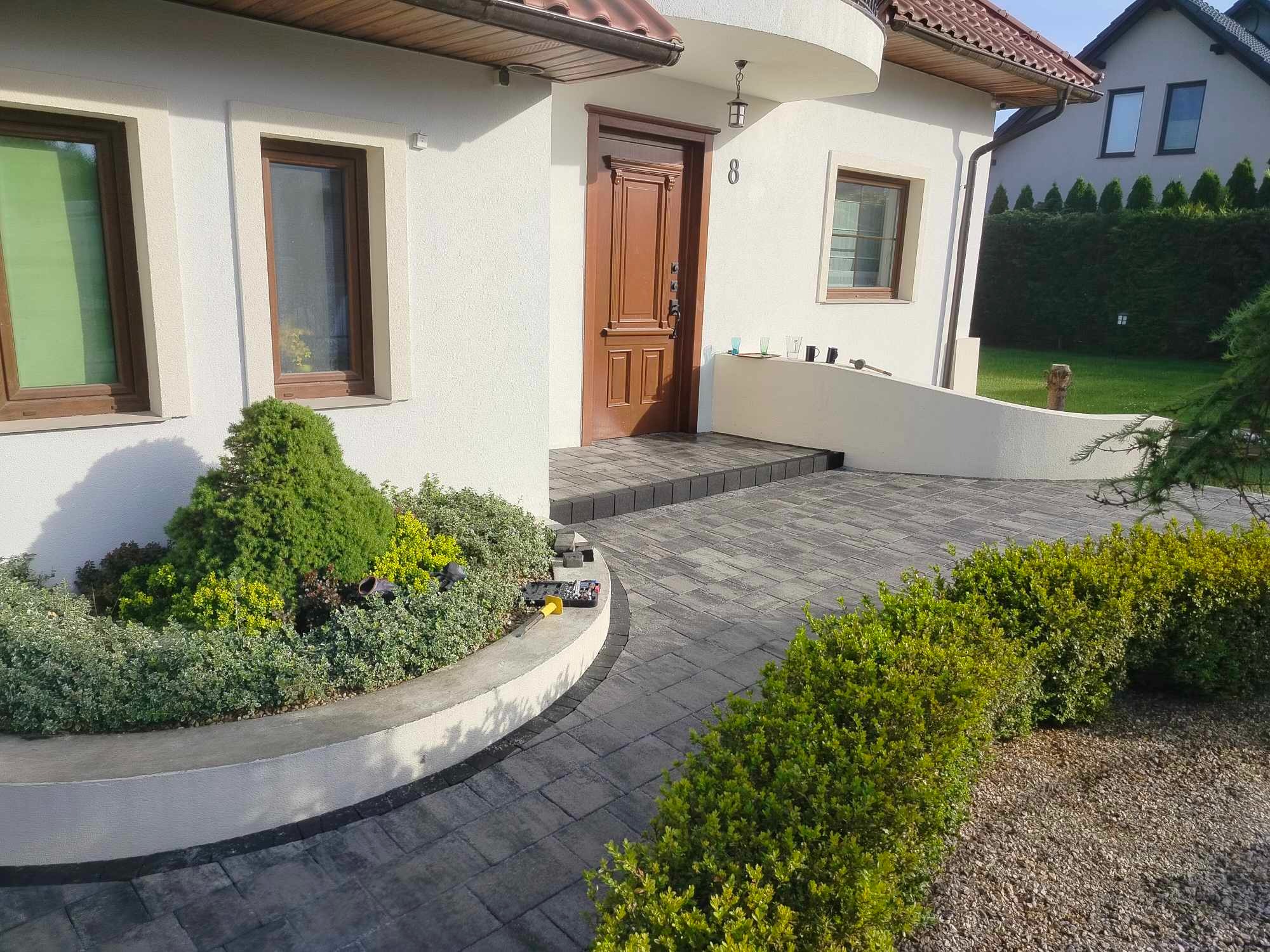 Nowoczesny dom z jasną elewacją, ciemnobrązowymi oknami i drzwiami, numerem domu '8', oraz ścieżką z szarej kostki brukowej prowadzącą do wejścia, obok której znajduje się rabata z zielenią.
