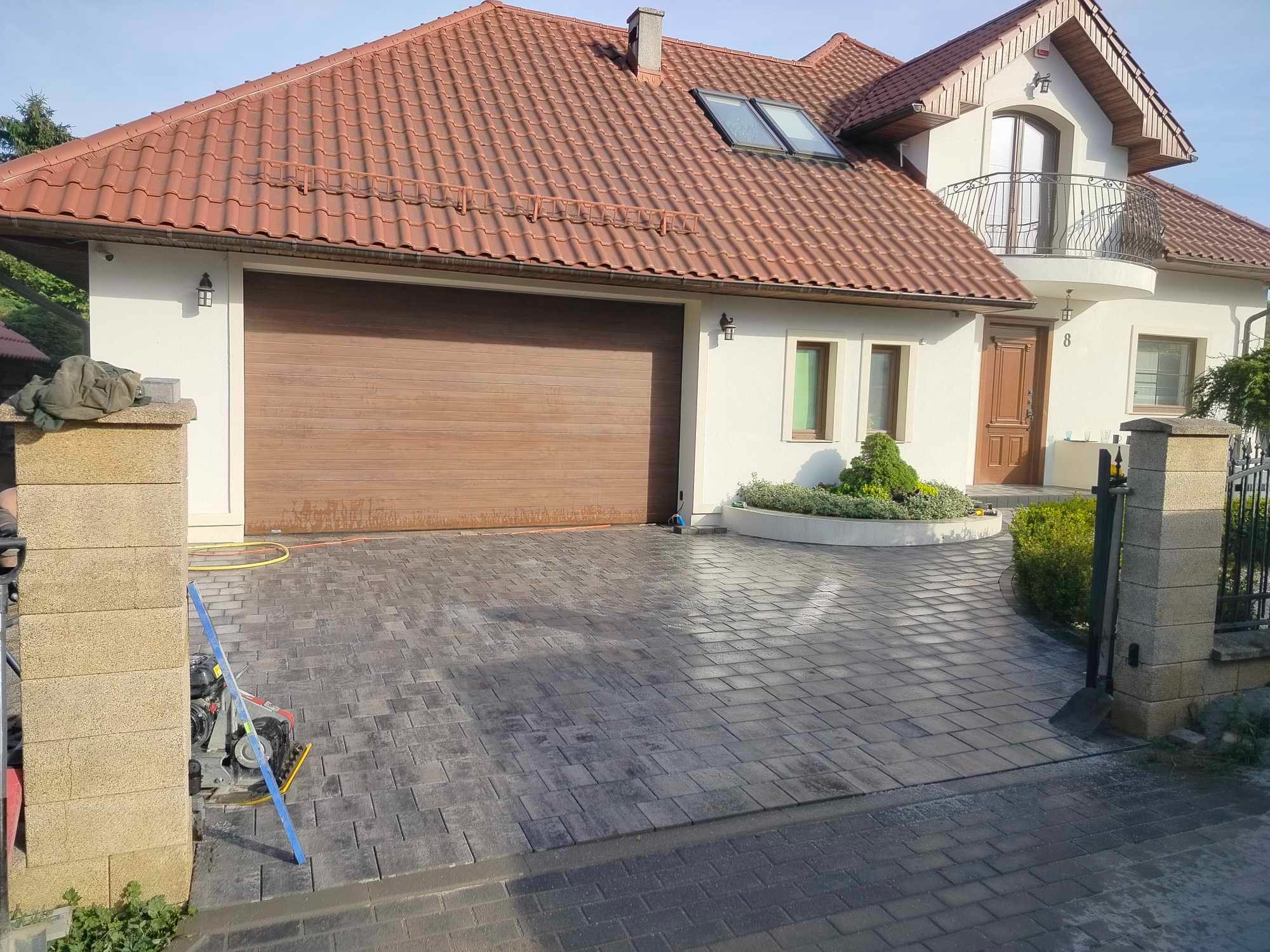 Nowo ułożona, szara kostka brukowa na podjeździe przed domem z brązową bramą garażową i czerwonym dachem, widoczna zagęszczarka i poziomica.