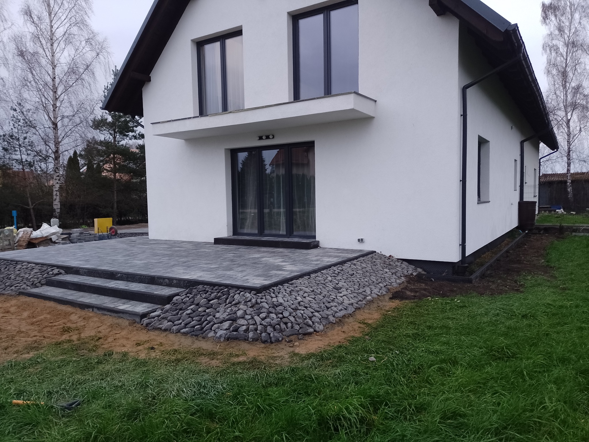 Nowoczesny taras z kostki brukowej w odcieniach szarości, otoczony dekoracyjnym kamieniem rzecznym, prowadzący do domu z białymi ścianami i czarnymi ramami okiennymi.