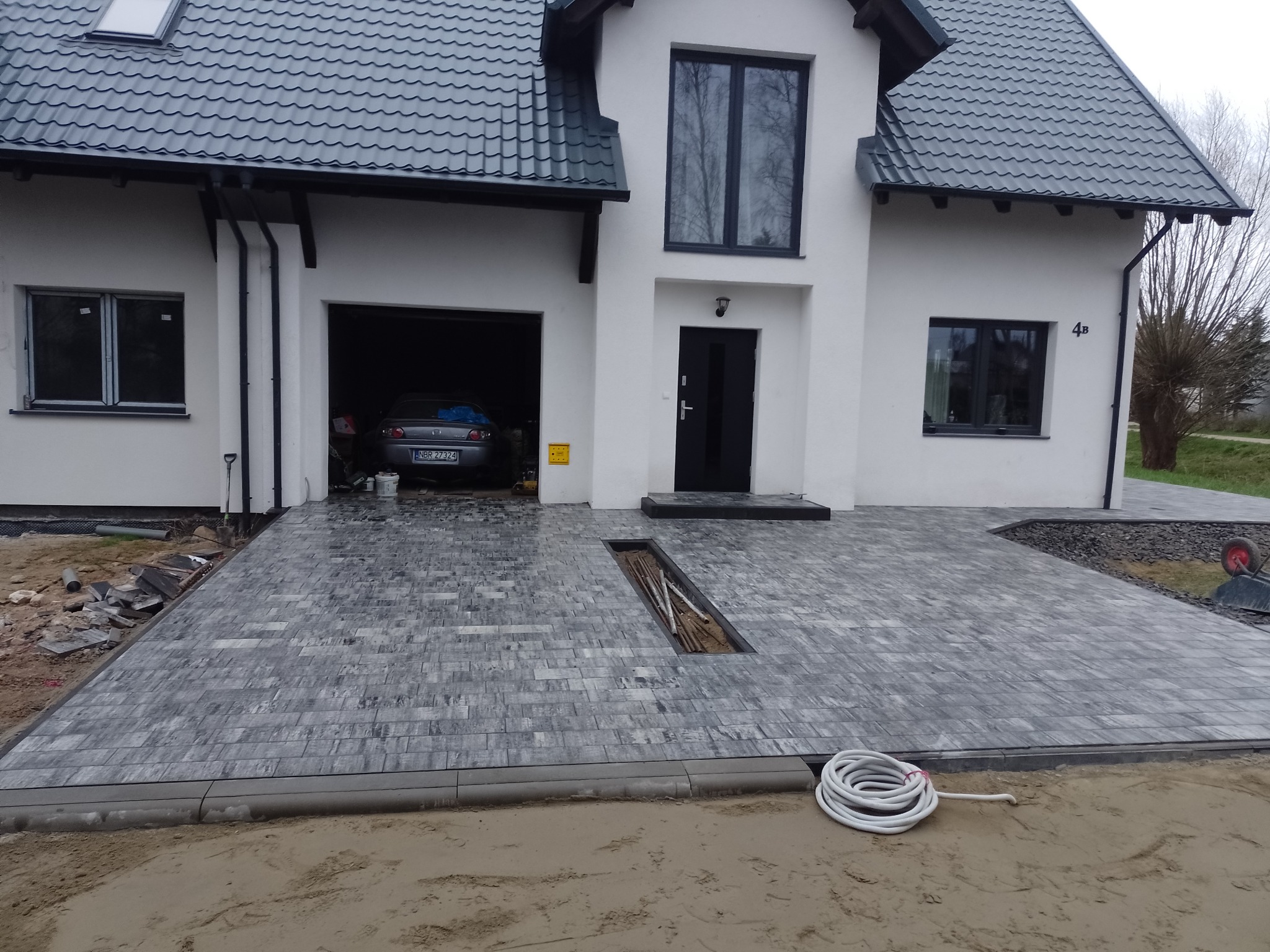 Nowo ułożona, szara kostka brukowa przed domem w Gdańsku, z widocznym odpływem liniowym i częściowo wykończonym obrzeżem. W tle garaż z zaparkowanym samochodem.