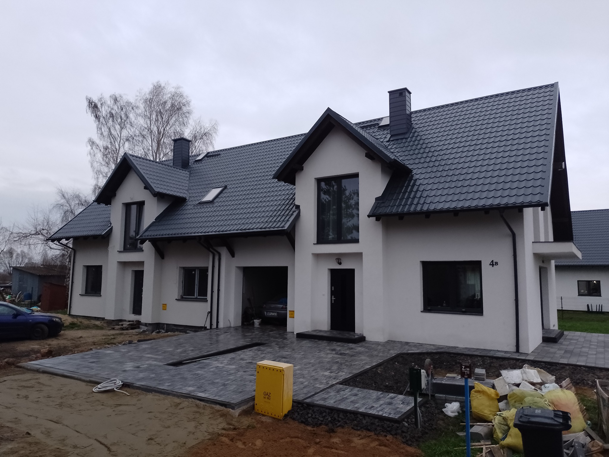 Nowoczesny szeregowiec z szarą kostką brukową, widoczny żółty licznik gazu i częściowo zagospodarowany teren wokół domu.