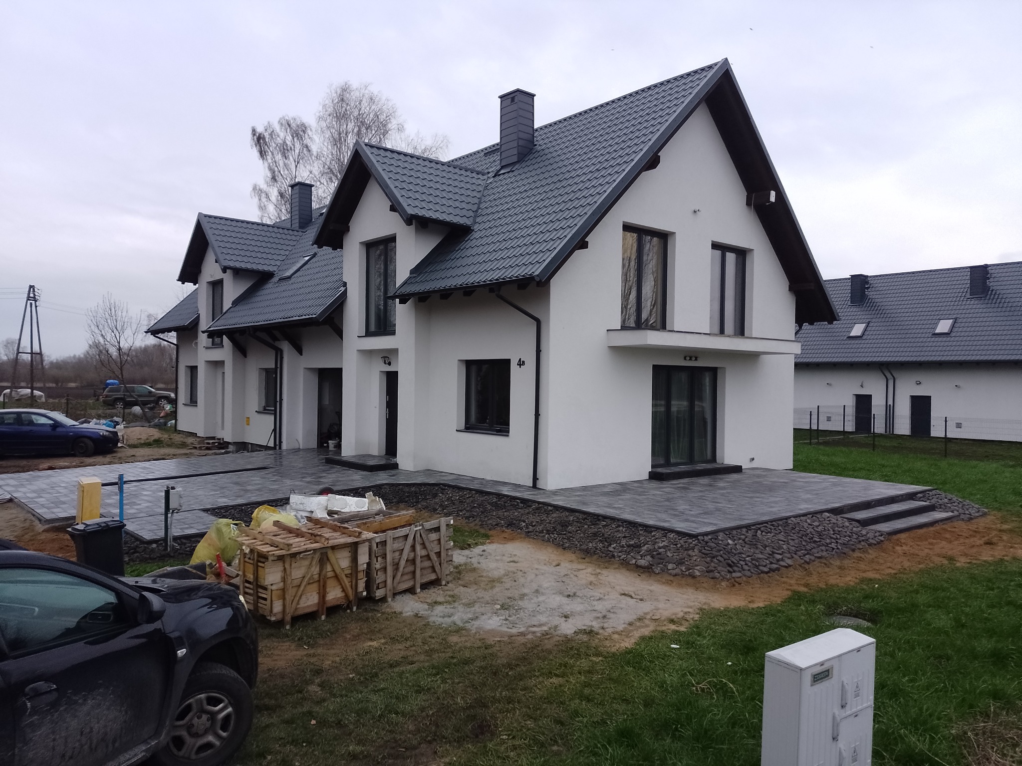 Nowoczesne szeregówki z szarej kostki brukowej na tarasach i podjazdach, widoczne drewniane skrzynie z materiałami budowlanymi, samochód osobowy i fragment trawnika.