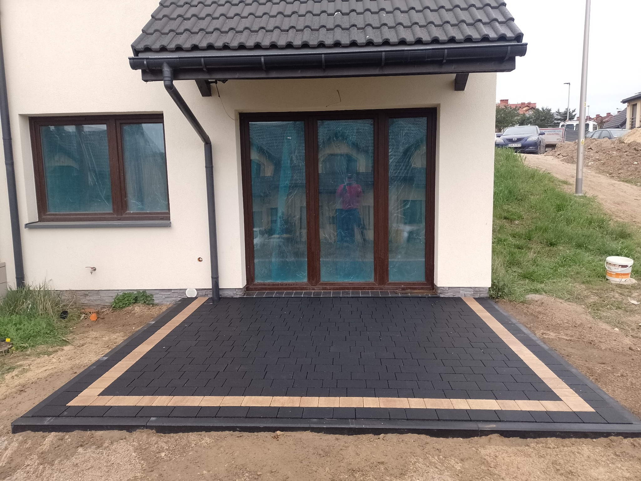 Nowo ułożony taras z ciemnej kostki brukowej z obramowaniem z jasnej kostki, przylegający do ściany domu z brązowymi oknami i drzwiami tarasowymi, widoczny fragment ogrodu i wiadro.