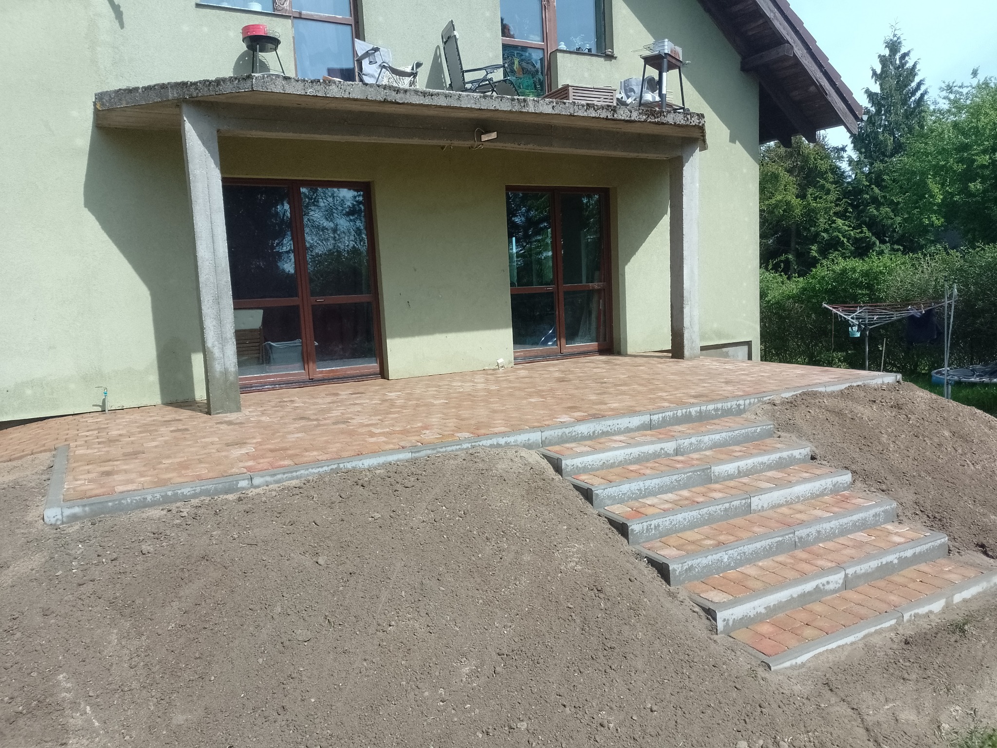 Wykończony taras z kostki brukowej z nowo wybudowanymi schodami prowadzącymi do ogrodu, z widocznym nasypem ziemi po bokach.