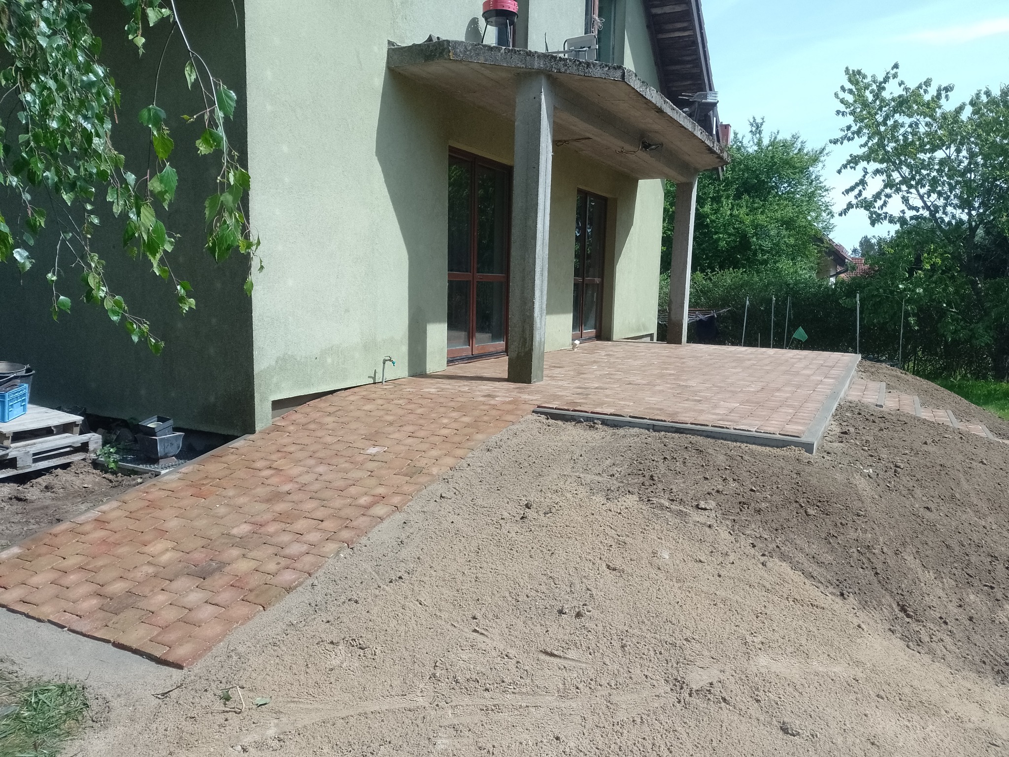 Ułożona ścieżka z czerwonej kostki brukowej prowadząca do tarasu przed domem, z widocznym nasypem ziemi po bokach.